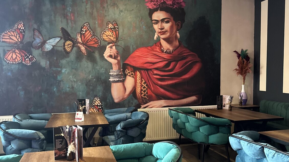 Ein stilvolles Café mit gemütlichen blauen Stühlen und Tischen. An der Wand ist ein großes Kunstwerk einer Frau mit Schmetterlingen. | © Bonbon