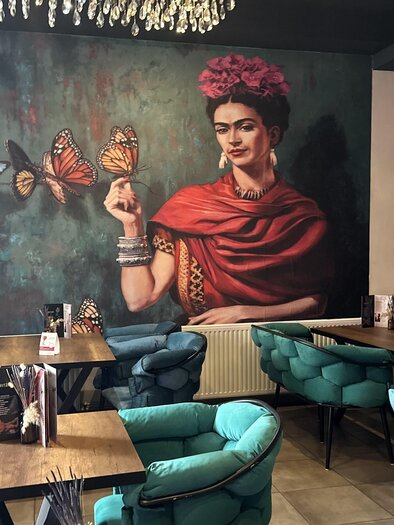 A stylish café with cozy blue chairs and tables.  
On the wall is a large artwork of a woman with butterflies. | © Bonbon