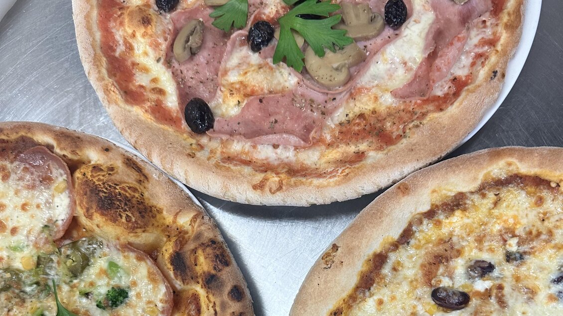 Drei köstliche Pizzen auf einem Tisch. Jede Pizza ist mit verschiedenen Zutaten belegt, darunter Schinken, Oliven und frische Kräuter. | © Bonbon