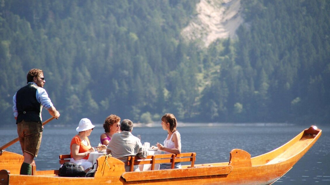 Eine Gruppe von fünf Personen sitzt in einem Holzboot auf einem ruhigen See. Im Hintergrund sind grüne Wälder und ein Hang zu sehen. | ©  Boote Salzkammergut