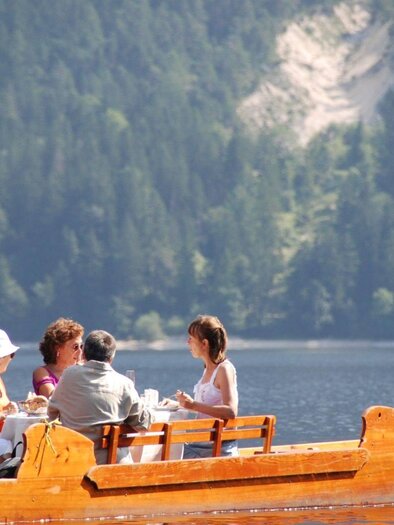 A group of five people sits in a wooden boat on a calm lake. In the background, green forests and a hillside can be seen. | ©  Boote Salzkammergut
