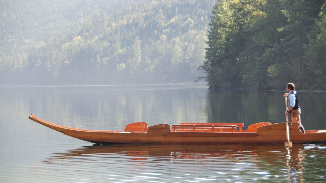 Ein Mann steht auf einem traditionellen Holzboot auf einem ruhigen See. Im Hintergrund sind grüne Wälder und sanfte Hügel zu sehen. | © Boote Salzkammergut