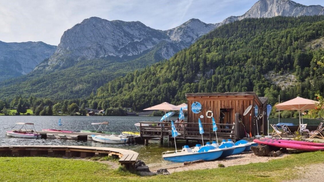 Ein malerischer See mit Booten und einer Holzhütte. Im Hintergrund sind majestätische Berge und grüne Wälder zu sehen. | © Bettina Scheck