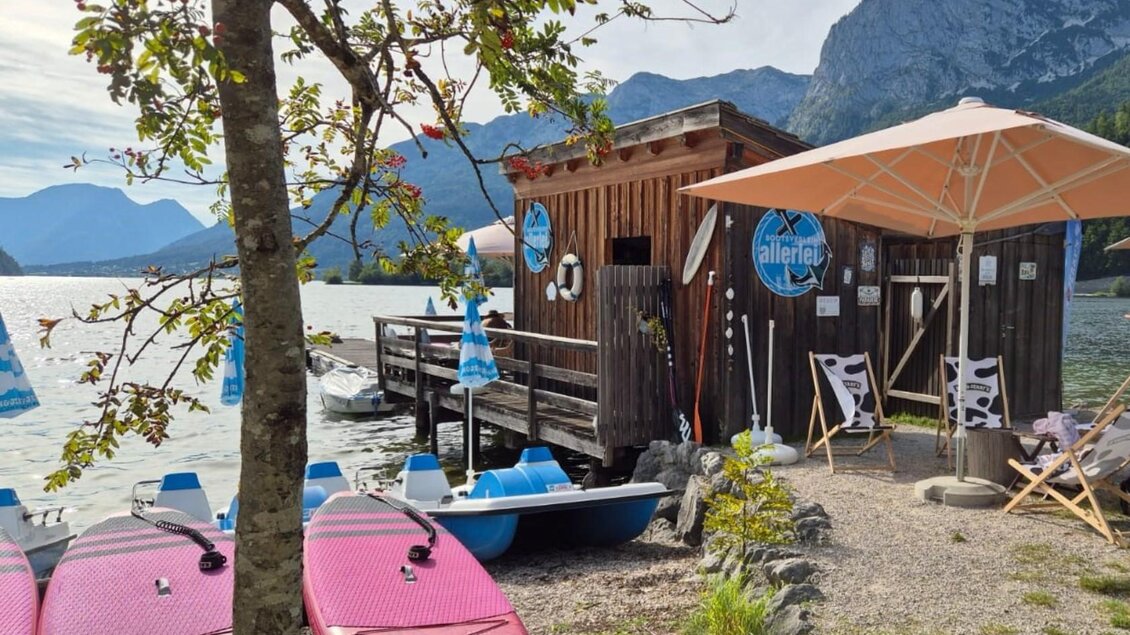 Ein malerischer Ort am Wasser mit einem Holzhaus und komfortablen Liegestühlen. Im Vordergrund stehen Kajaks bereit, um das Wasser zu erkunden. | © Bettina Scheck