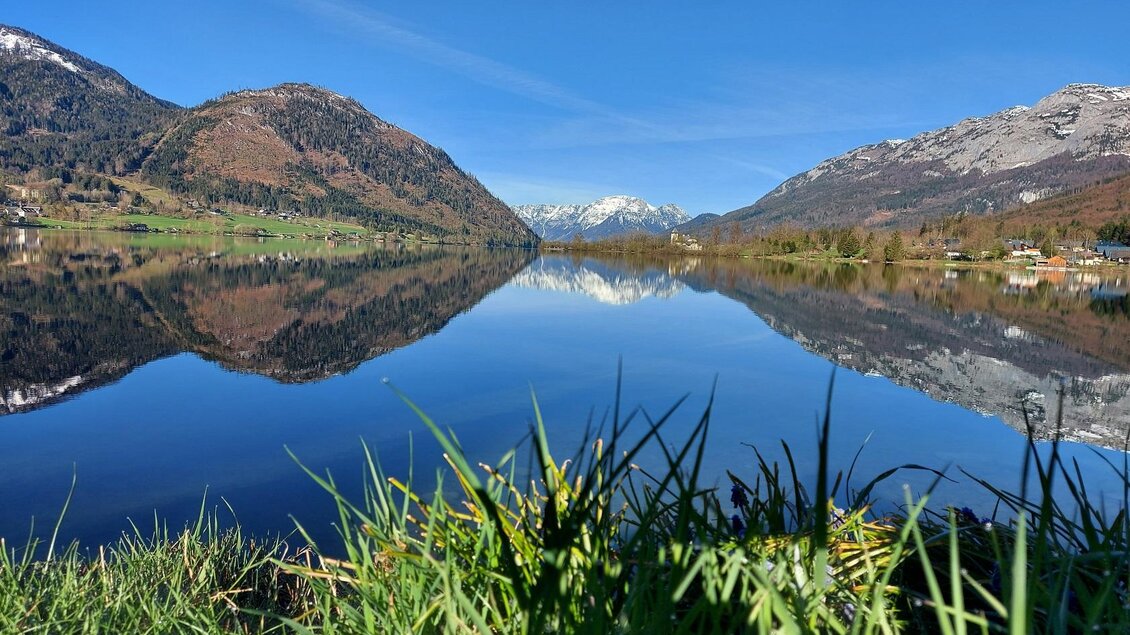 Ein ruhiger See, umgeben von Bergen und grünem Gras im Vordergrund. Der klare Himmel spiegelt sich perfekt im Wasser. | © Steinegger