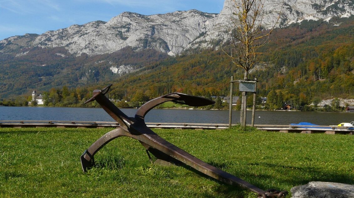 Ein Anker liegt auf einer Wiese am Ufer eines Sees. Im Hintergrund sind Berge und klare, blaue Himmel zu sehen. | © Rostiger Anker