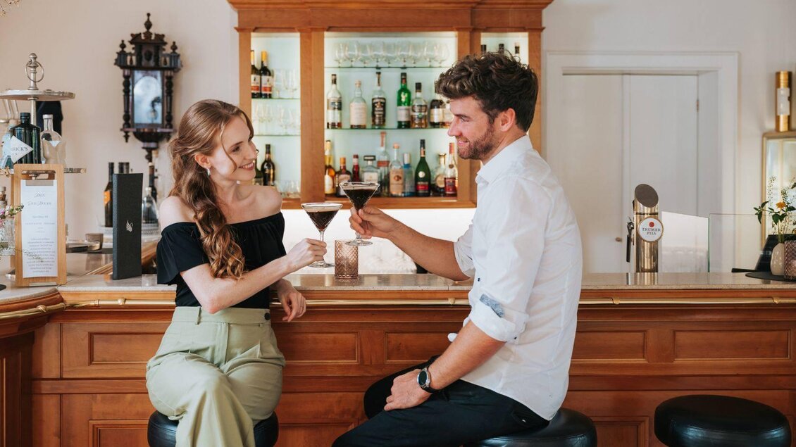 Ein Paar sitzt an einer Bar und stößt mit Cocktails an. Der Hintergrund zeigt eine gut sortierte Bar mit Getränken. | © Karl Steinegger