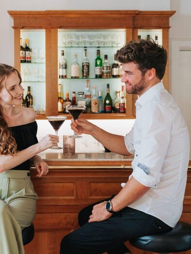 A couple is sitting at a bar, clinking their cocktails together. The background features a well-stocked bar with drinks. | © Karl Steinegger