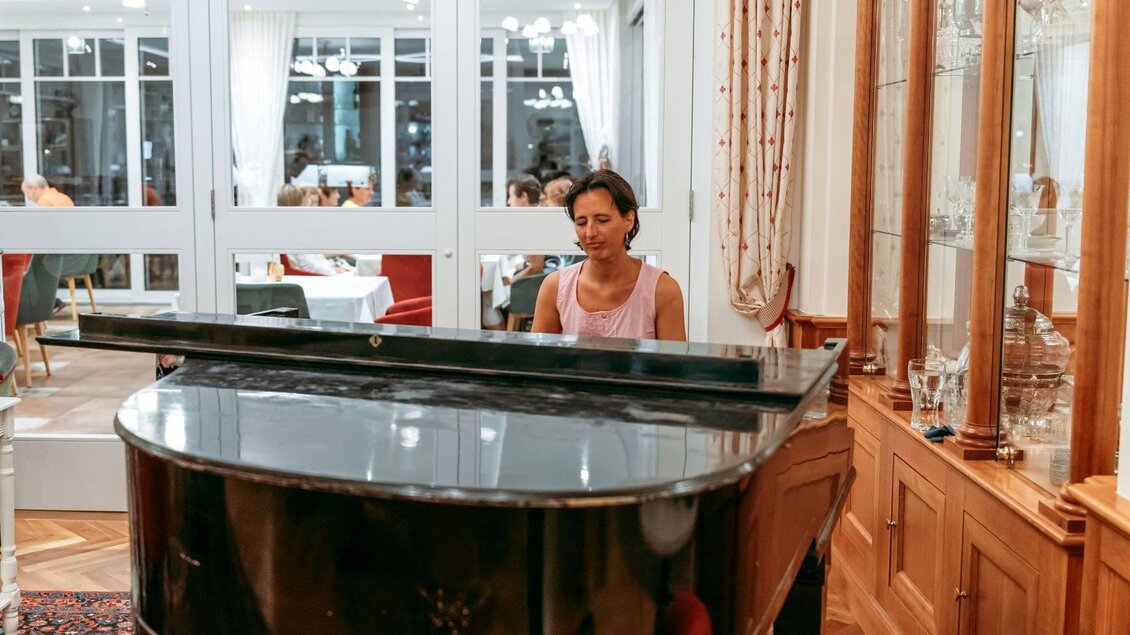 Eine Frau spielt Klavier in einem eleganten Raum. Im Hintergrund sind Tische und Gäste in einem Restaurant zu sehen. | © Karl Steinegger