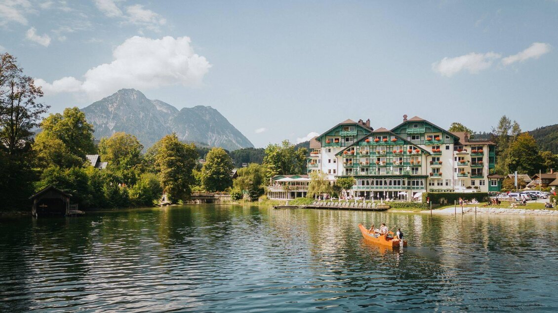 Ein malerisches Hotel am Ufer eines ruhigen Sees, umgeben von Bergen und Bäumen. Zwei Personen paddeln in einem orangefarbenen Boot auf dem Wasser. | © Karl Steinegger