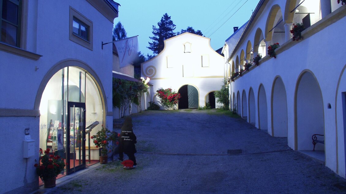 Eine ruhige Straße mit weißen Gebäuden im Abendlicht. Rechts sind Bögen und blühende Pflanzen zu sehen. | © Brahmsmuseum