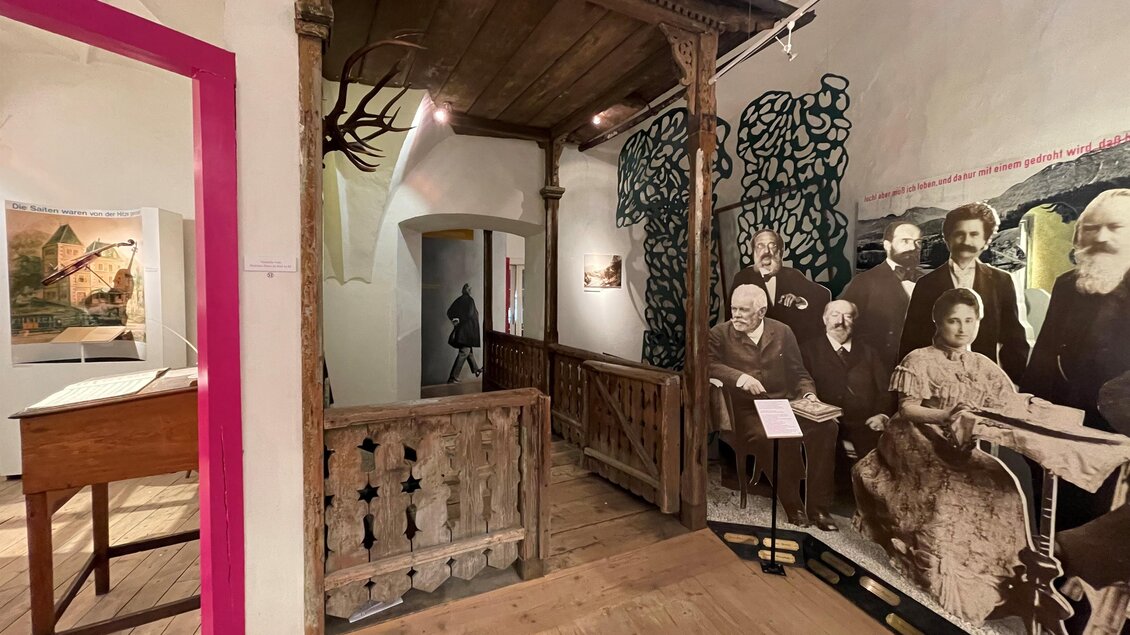 Ein historischer Innenraum mit Holzelementen und einer Wand, die Schwarz-Weiß-Fotos von Menschen zeigt. Im Hintergrund sind Kunstwerke und Informationen zu sehen. | © Brahmsmuseum