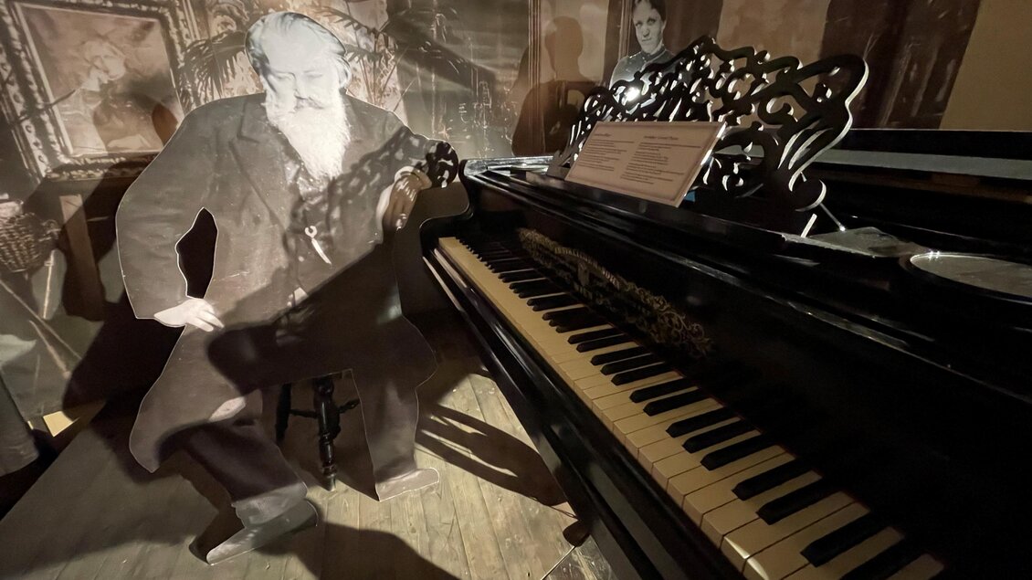 Ein historisches Klavier steht im Vordergrund, neben einer stilisierten Figur eines Mannes. Im Hintergrund sind Schattenspiele und Bilder zu sehen, die eine nostalgische Atmosphäre erzeugen. | © Brahmsmuseum