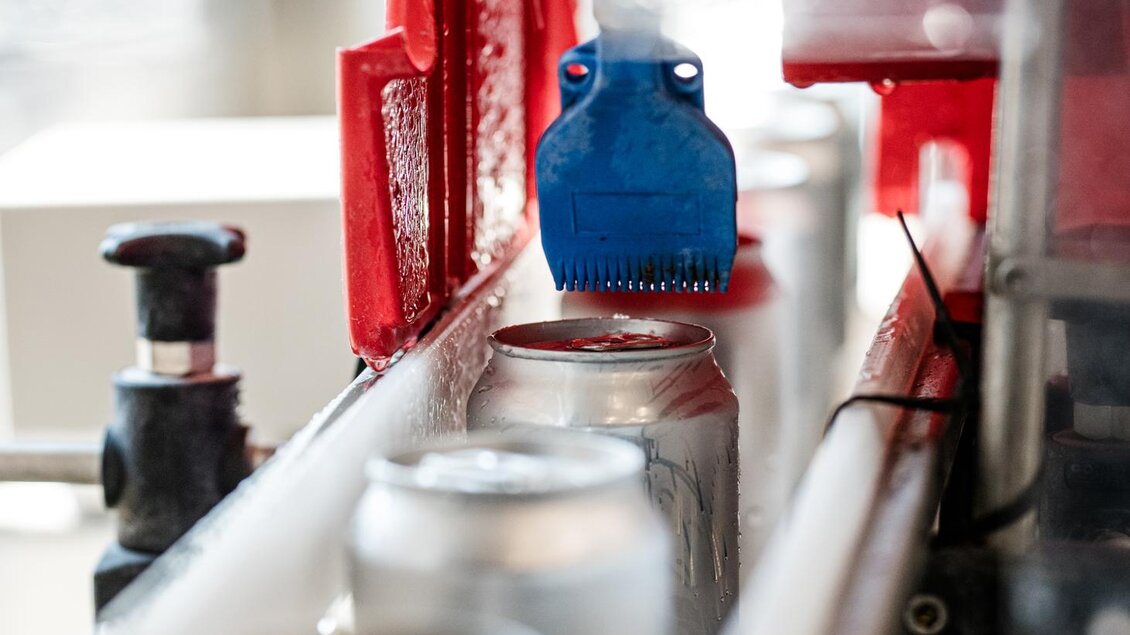 Eine moderne Abfüllanlage mit Dosen, die auf einem Förderband transportiert werden. Ein blauer Pinsel reinigt die Dosen, während sie durch den Produktionsprozess gehen. | © Brauhaus Bevog