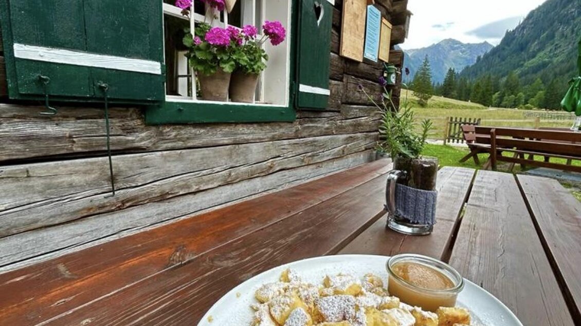 Ein Teller mit süßen Knödeln, bestäubt mit Puderzucker, steht auf einem Holztisch. Im Hintergrund sieht man eine rustikale Holzhütte und die malerische Berglandschaft. | © Breitlahnhütte