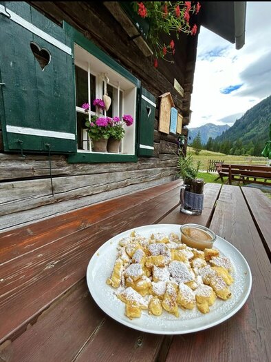 A plate of sweet dumplings, dusted with powdered sugar, is sitting on a wooden table. In the background, you can see a rustic wooden cabin and the picturesque mountain landscape. | © Breitlahnhütte