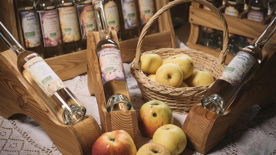Ein Korb mit Äpfeln und Flaschen äpfeligem Getränk stehen auf einem Tisch. Im Hintergrund sind weitere Flaschen sichtbar, die in Holzregalen platziert sind. | © Stefan Leitner