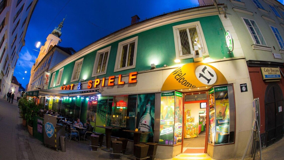 Ein belebtes Lokal mit bunten Lichtern und einem einladenden Eingang. Die Fassade ist grün und orange, und es sind Tische im Freien sichtbar. | © Brot & Spiele