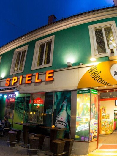 A lively establishment with colorful lights and an inviting entrance. The facade is green and orange, and outdoor tables are visible. | © Brot & Spiele