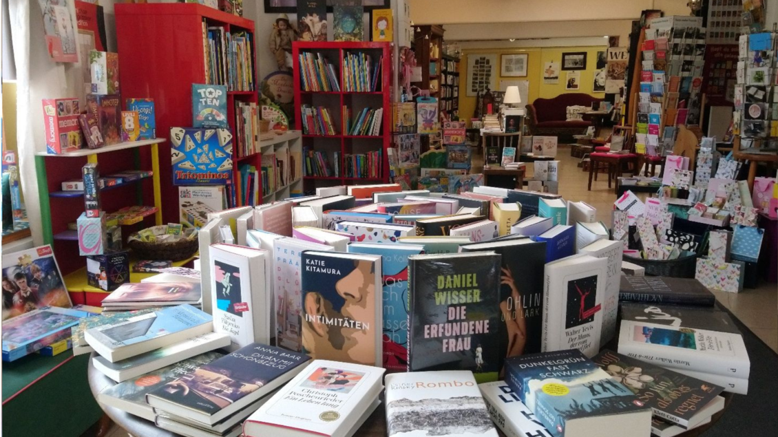 Ein gemütlicher Buchladen mit vielen bunt gestalteten Regalen. In der Mitte steht ein runder Tisch, auf dem zahlreiche Bücher ausgestellt sind.