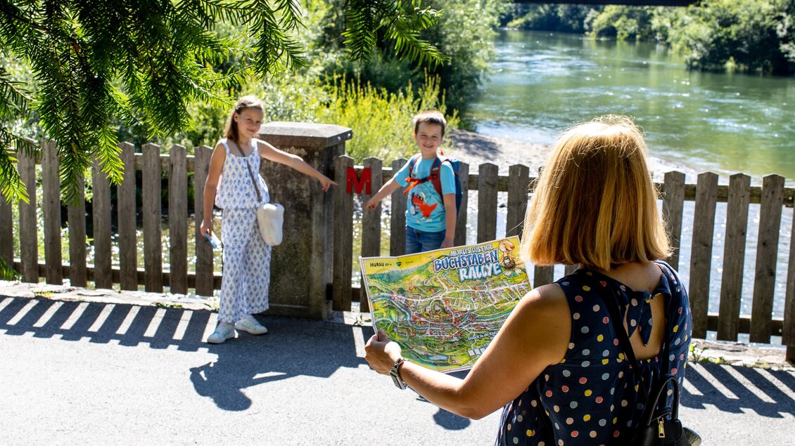 Eine Frau zeigt einer Gruppe von Kindern eine Karte. Im Hintergrund fließt ein ruhiger Fluss und es gibt Bäume. | © TVB Murau