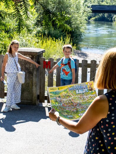 A woman is showing a group of children a map. In the background, a calm river flows and there are trees. | © TVB Murau