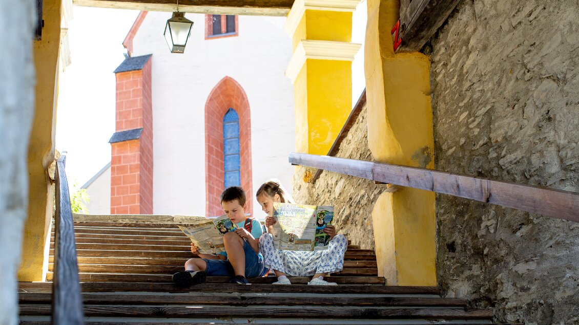 Zwei Kinder sitzen auf einer Treppe und lesen Karten. Im Hintergrund ist ein Gebäude mit roter und gelber Fassade zu sehen. | © TVB Murau