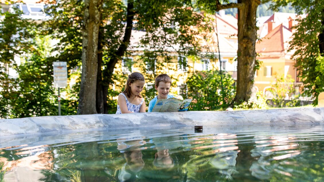 Zwei Kinder sitzen am Rand eines Wasserspiels und schauen in ein Buch. Im Hintergrund sind grüne Bäume und farbige Gebäude zu sehen. | © TVB Murau