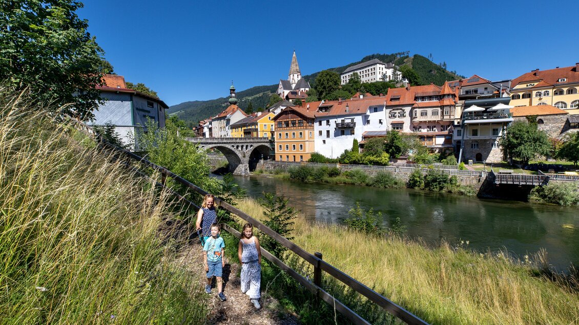 Eine malerische Stadtansicht mit bunten Häusern am Fluss. Im Vordergrund spazieren drei Kinder auf einem Weg. | © TVB Murau