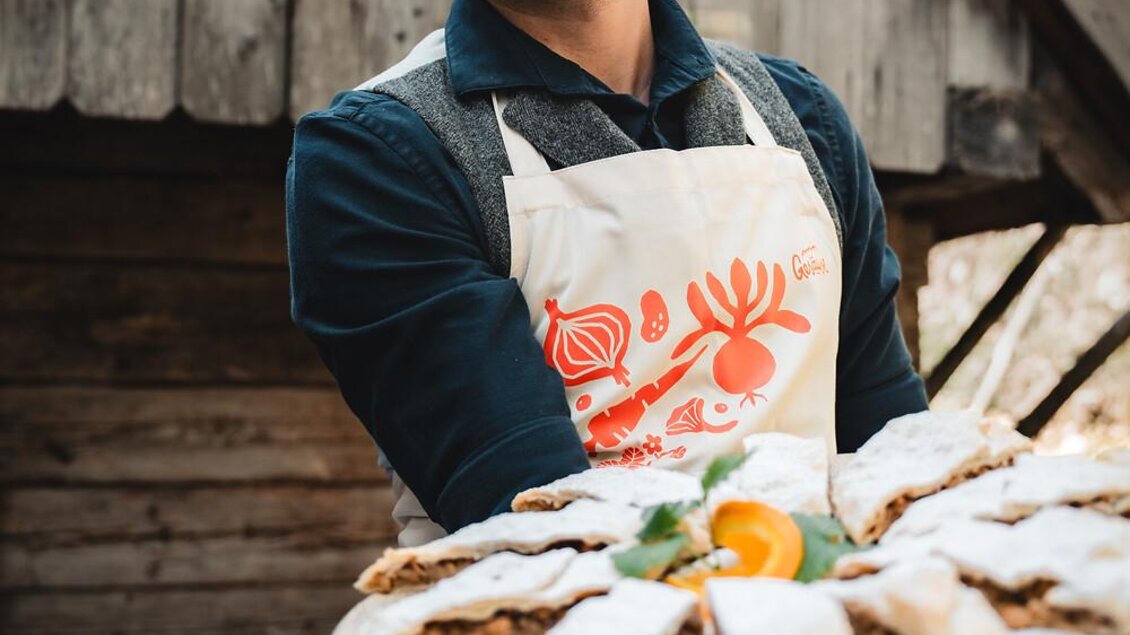 Ein Mann trägt eine große Platte mit Gebäck und lächelt. Im Hintergrund ist eine rustikale Holzstruktur zu sehen.