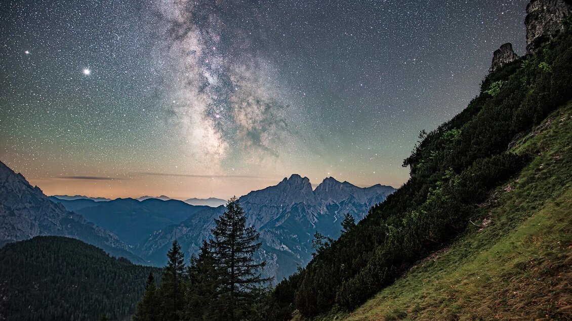 Ein beeindruckender Nachthimmel mit der Milchstraße und funkelnden Sternen. Im Vordergrund sind majestätische Berge und grüne Wälder zu sehen.