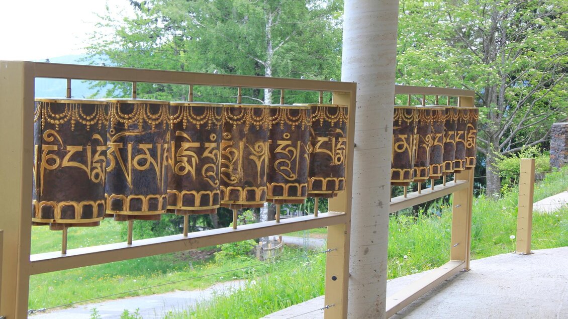 Eine Reihe von großen, goldenen Gebetsfahnen an einem Holzgestell. Im Hintergrund sind grüne Bäume und eine ruhige Landschaft zu sehen. | © TV Sulmtal Koralm