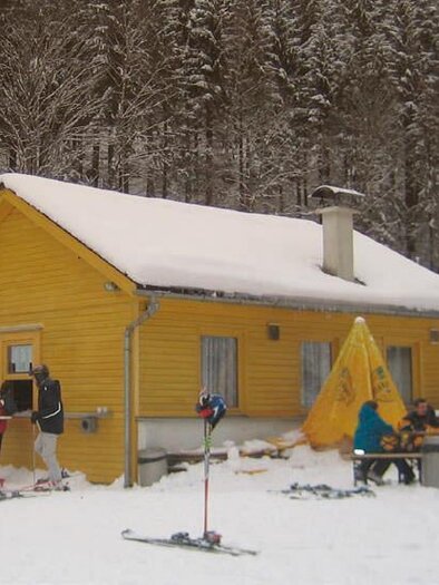 A yellow mountain cabin surrounded by snow and forest. People stand with ski equipment and sit at tables outside. | © Berglift
