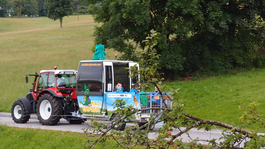 Ein Traktor zieht eine kleine Fahrerhaube auf einer Landstraße. Im Hintergrund sind grüne Felder und Bäume zu sehen. | © Sattelberghütte