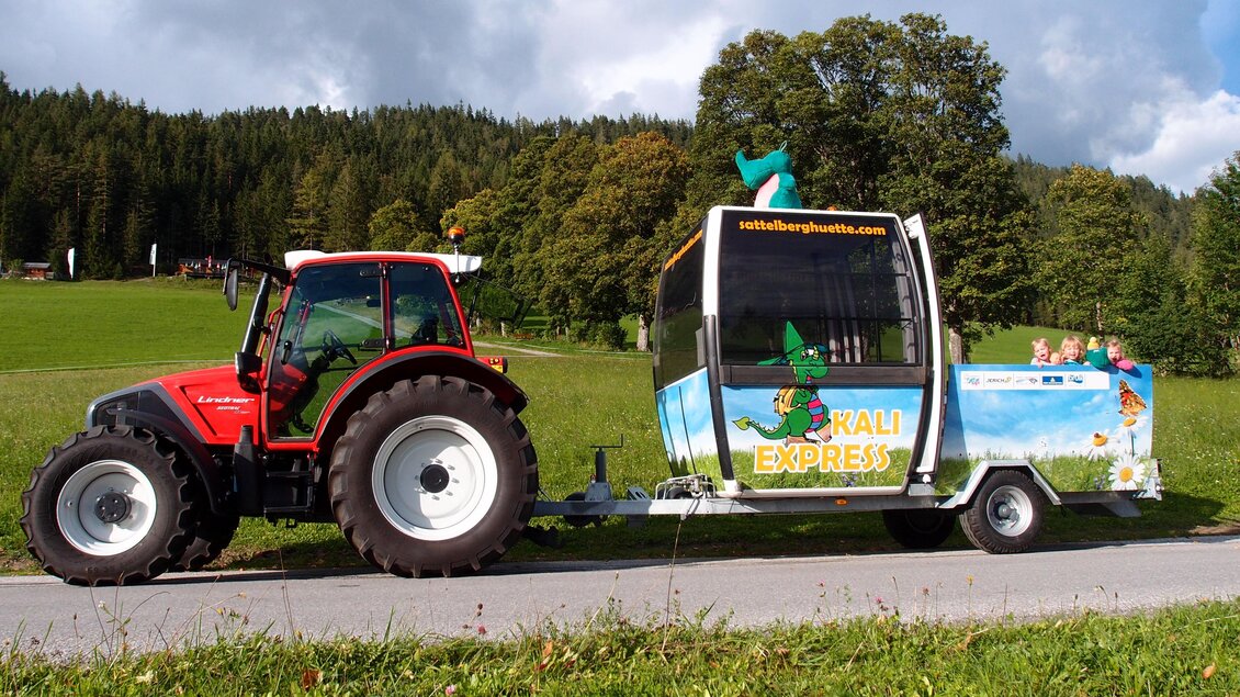 Ein roter Traktor zieht einen bunten Anhänger mit Werbung. Im Hintergrund sind grüne Wiesen und Bäume zu sehen. | © Sattelberghütte