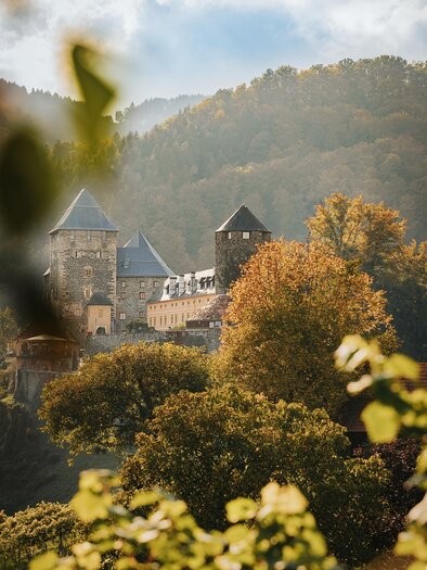 Eine malerische Burg umgeben von buntem Laub und sanften Hügeln. Der Himmel ist klar und verleiht der Szenerie eine friedliche Atmosphäre.