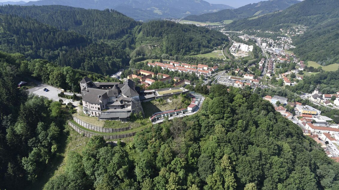 Eine beeindruckende Burg liegt auf einem Hügel, umgeben von grünen Wäldern. Im Hintergrund sind sanfte Berge und eine kleine Stadt zu sehen. | © Stadtgemeinde Kapfenberg