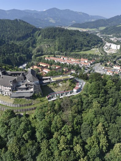 An impressive castle sits on a hill, surrounded by green forests. In the background, gentle mountains and a small town can be seen. | © Stadtgemeinde Kapfenberg