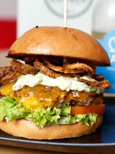 A delicious burger with melted cheese, fresh vegetables, and crispy onion rings. In the background, you can see a drink and an elegant glass. | © Burger Factory