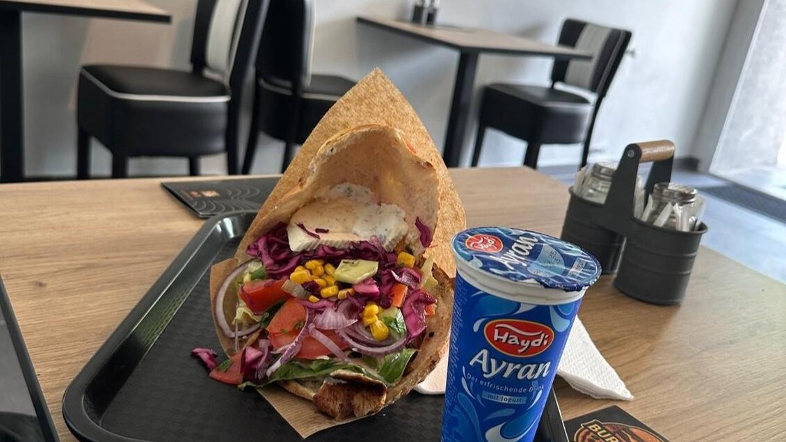 Ein Teller mit Döner im Fladenbrot, gefüllt mit buntem Gemüse. Daneben steht ein Becher Ayran auf einem Tisch in einem modernen Restaurant. | © Kevin Dönmez