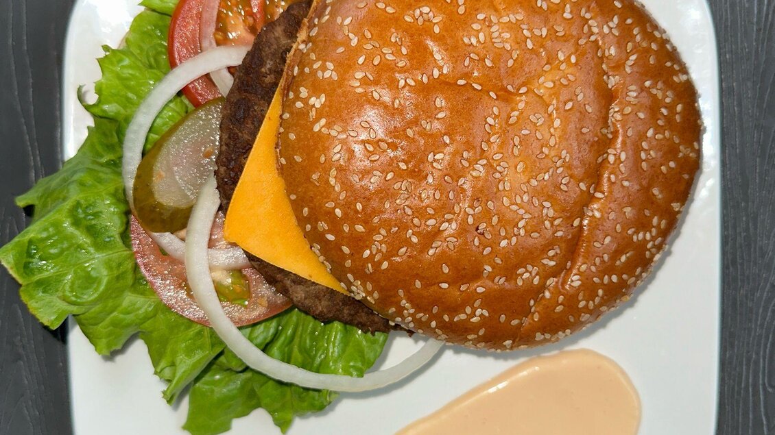 Ein saftiger Burger auf einem Teller, belegt mit Käse, frischem Gemüse und Salat. Daneben befindet sich eine Portion Sauce. | © Kevin Dönmez