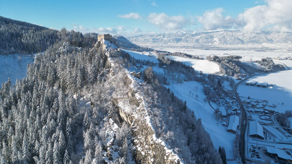 Eine schneebedeckte Hügelkette mit Bäumen und einem alten Turm. Im Hintergrund erstrecken sich die Berge unter einem strahlend blauen Himmel. | © Isabella Painhapp