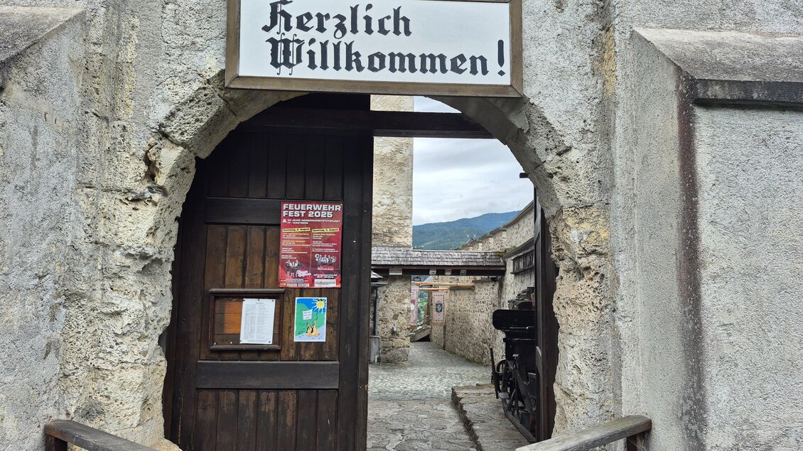 Ein historischer Eingang mit einem Schild, das "Herzlich Willkommen!" sagt. Dahinter ist eine steinerne Mauer und ein schöner Ausblick auf die Berge. | © Andrea Stelzer