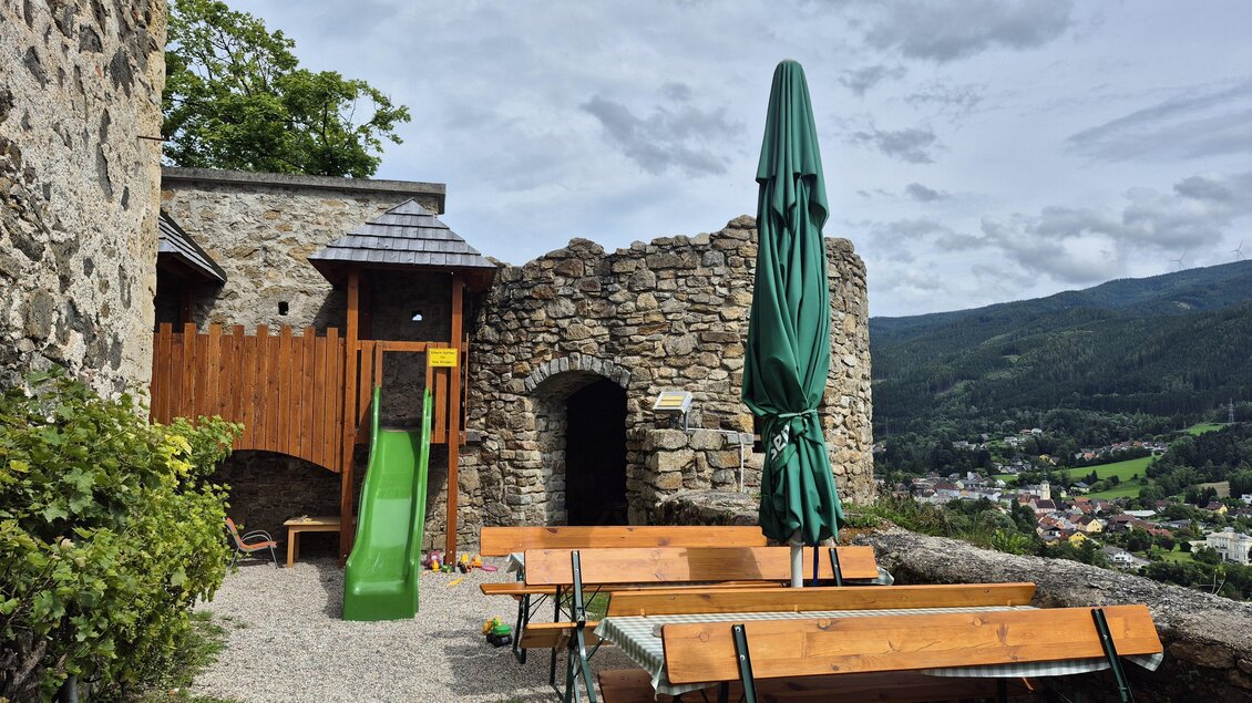 Ein Spielplatz mit einer Rutsche und Bänken befindet sich in der Umgebung einer alten Burg. Im Hintergrund ist die grüne Landschaft sichtbar. | © Andrea Stelzer