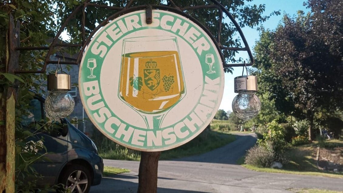 Ein Schild mit der Aufschrift "Steirischer Buschenschank" zeigt ein Glas Bier in der Mitte. Es ist von Bäumen umgeben und steht an einem Weg. | © Fam. Tomaschitz