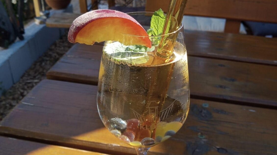 Ein erfrischendes Getränk in einem Weinglas, verziert mit einem Apfelschnitz und frischer Minze. Der Hintergrund zeigt ein Holztablett und Pflanzen. | © Fam. Tomaschitz