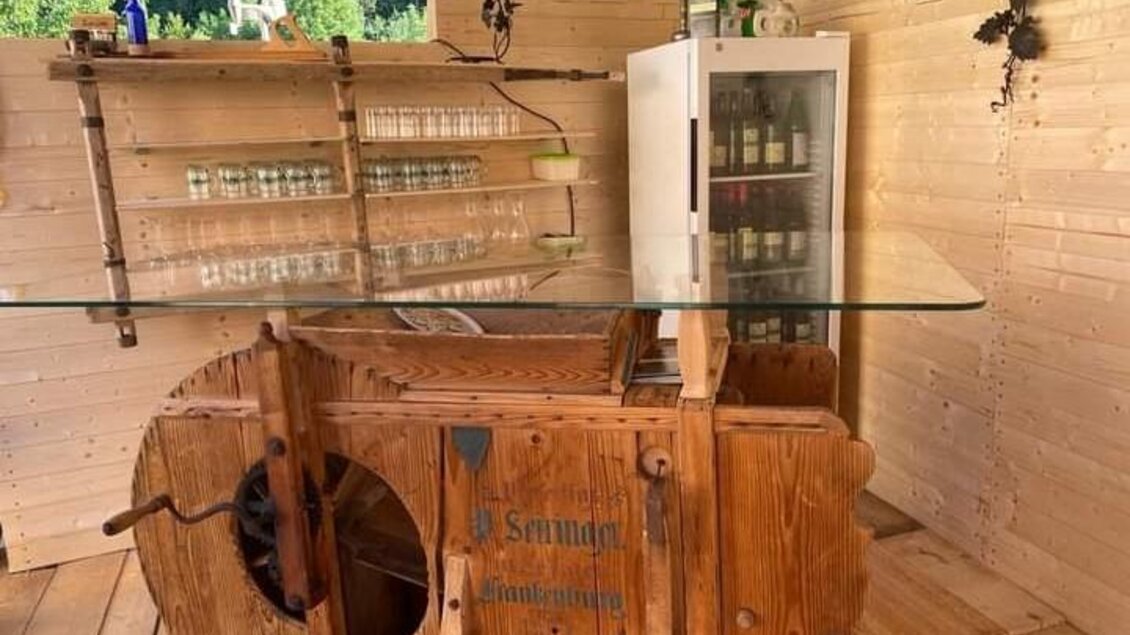 Eine rustikale Bar aus Holz mit einem Glastresen und Regalen für Gläser. Im Hintergrund steht ein Kühlschrank mit Getränken. | © Fam. Tomaschitz