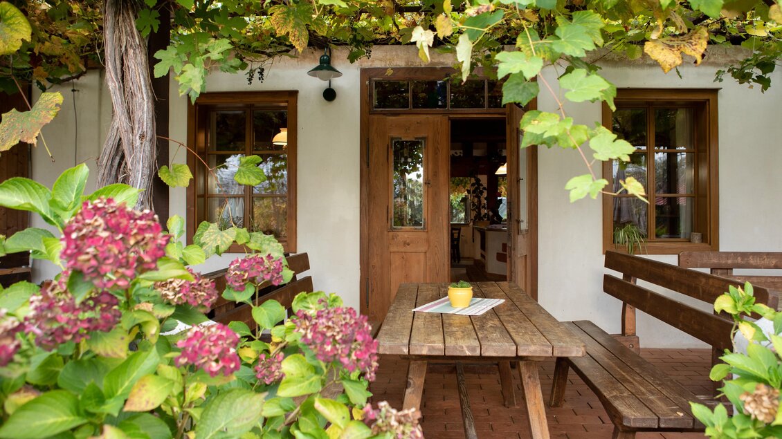 Ein gemütlicher Sitzbereich im Freien mit Holztisch und Bänken. Umgeben von bunten Blumen und Weintrauben. | © GlaMur-Genuss am Fluss