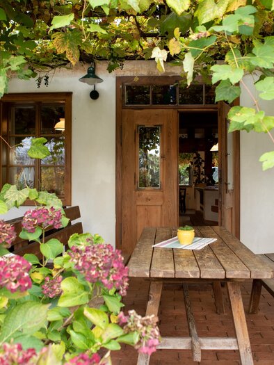 A cozy outdoor seating area with a wooden table and benches. Surrounded by colorful flowers and grapevines. | © GlaMur-Genuss am Fluss