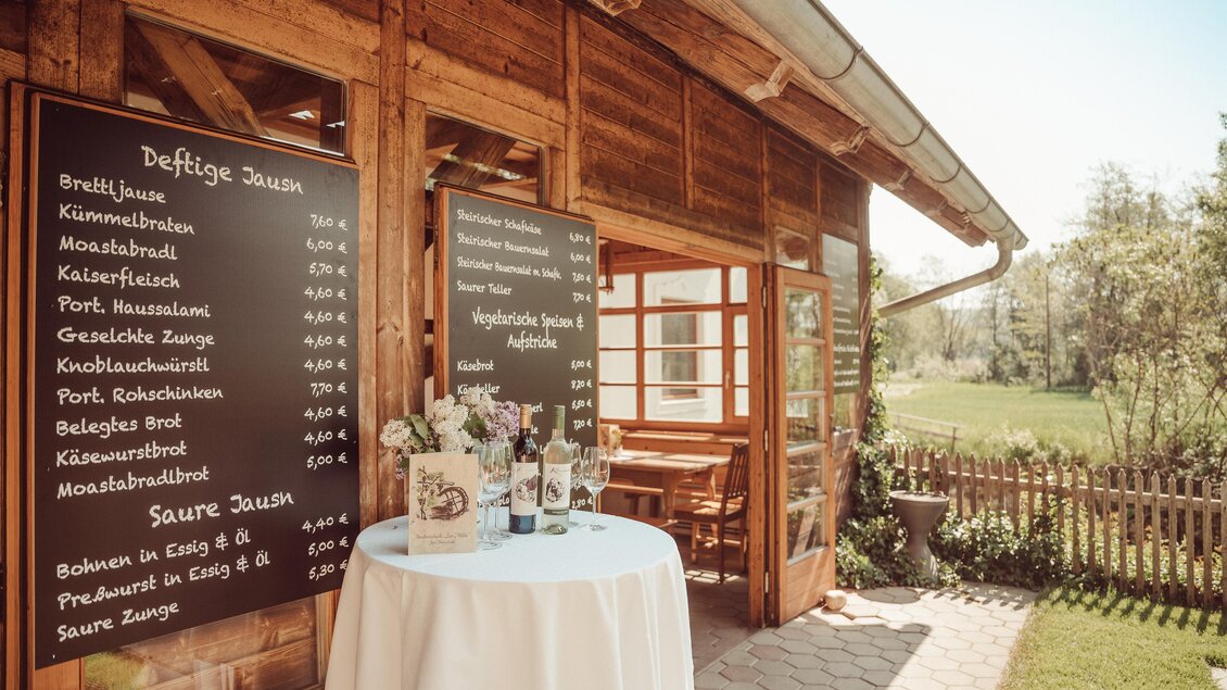 Ein einladendes Holzhäuschen mit einem Tisch und einer Blumenvasen-Dekoration. An der Wand hängen Tafeln mit Menüangeboten. | © Weingut Kleinschuster